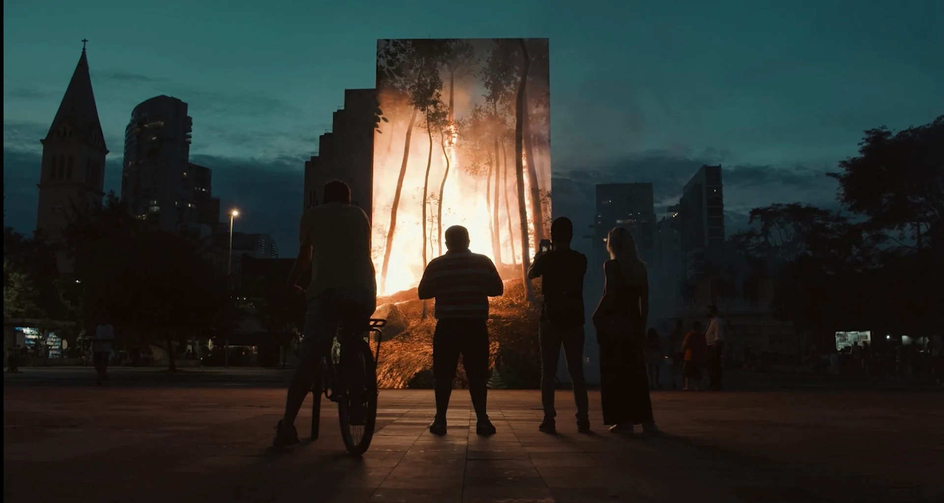 Visitors and a cyclist pause in front of MATA at dusk, with the illuminated forest centered against the skyline of Largo da Batata.